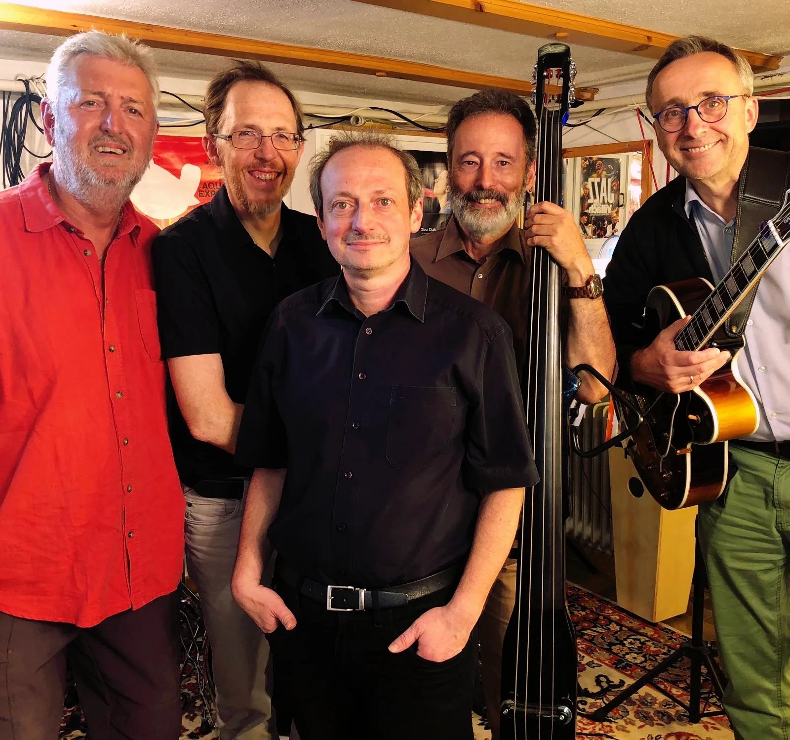 Five musicians standing in a rehearsal room with instruments.