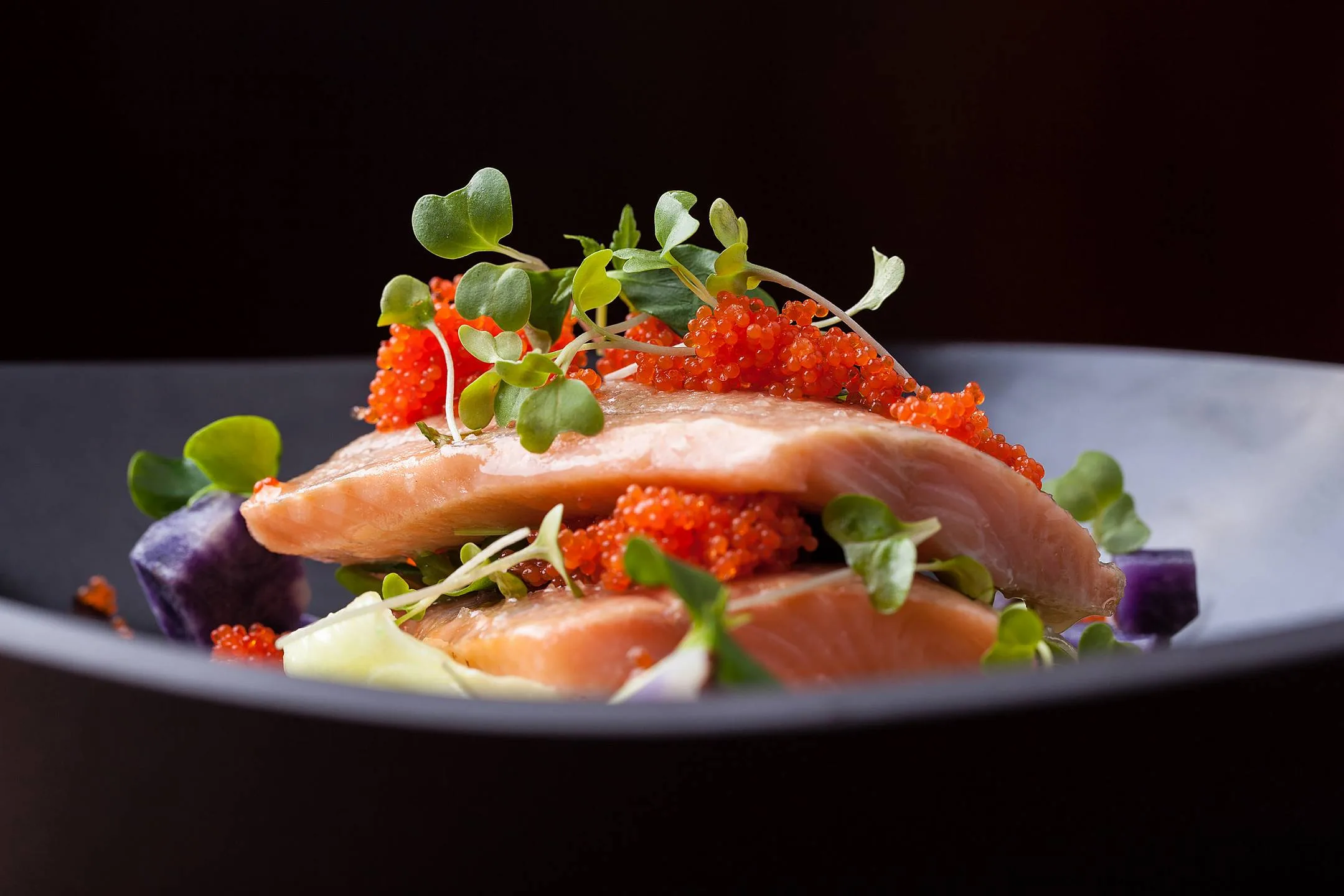 A dish featuring salmon, caviar, and fresh herbs in a black bowl.