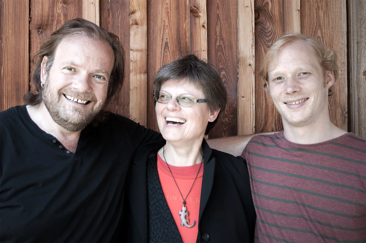 Three people smiling happily in front of a wooden wall.