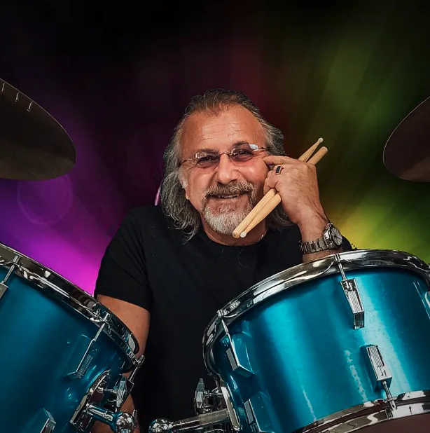 A smiling man sits behind a blue drum set holding drumsticks.