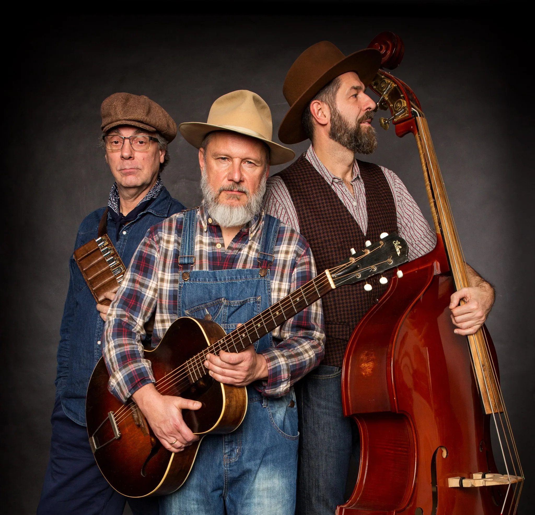 Three musicians stand together, one with a guitar, one with a double bass, and one with a harmonica.