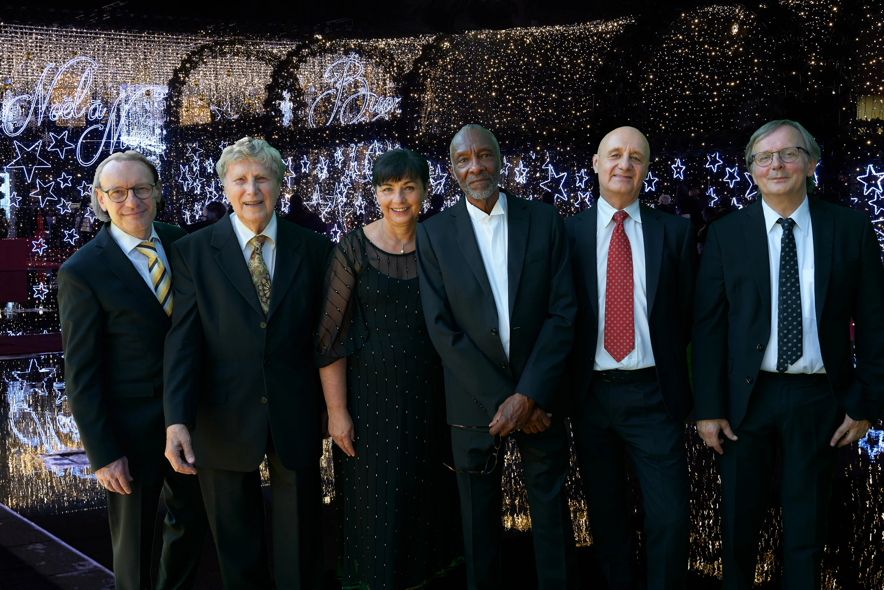 Six people in suits standing in front of a festive illuminated background.