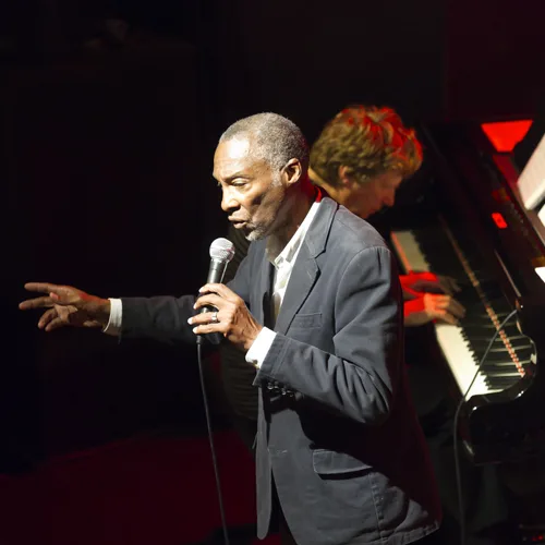 A singer with a microphone on stage, a pianist in the background.