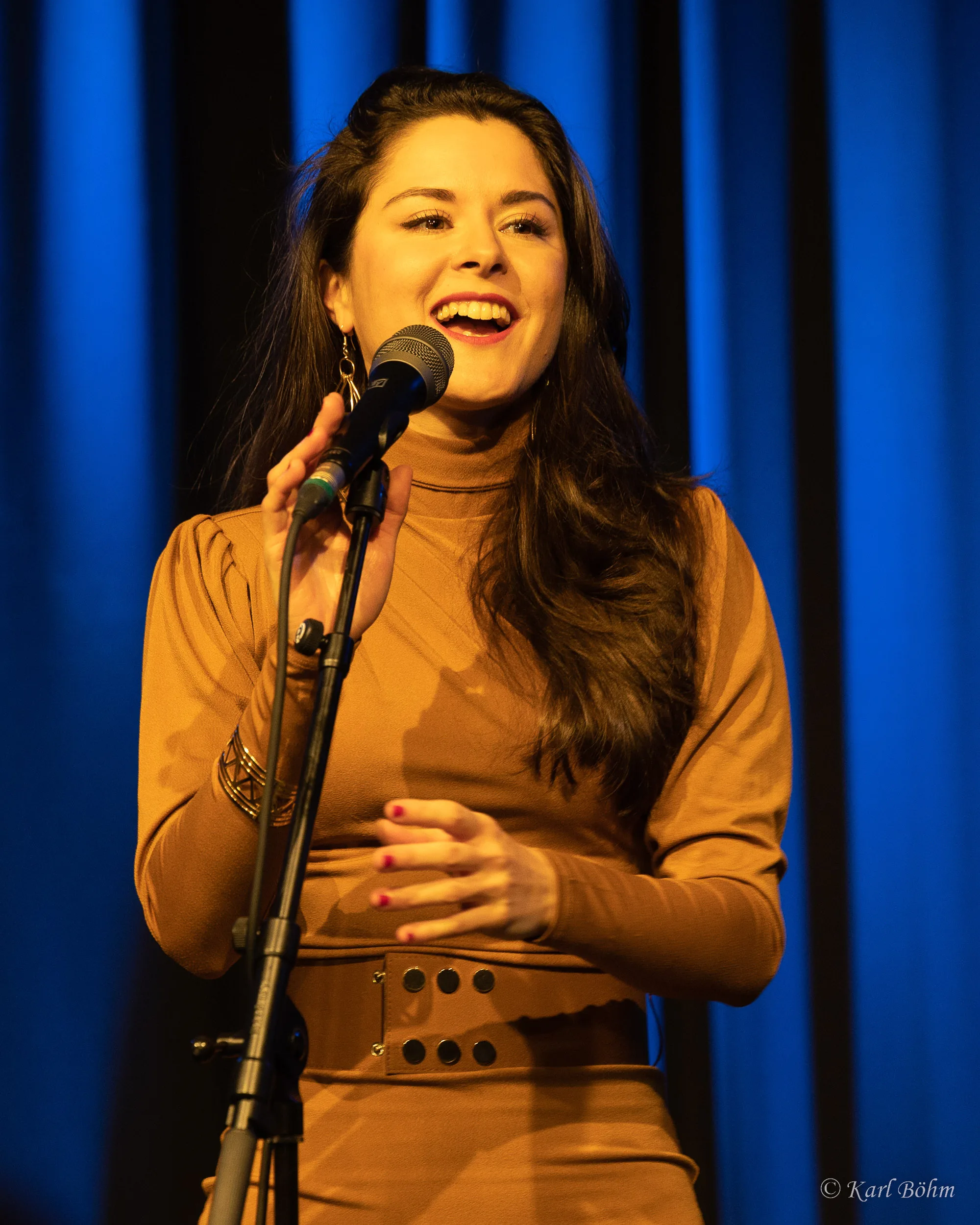A singer in a brown outfit sings into a microphone on stage.