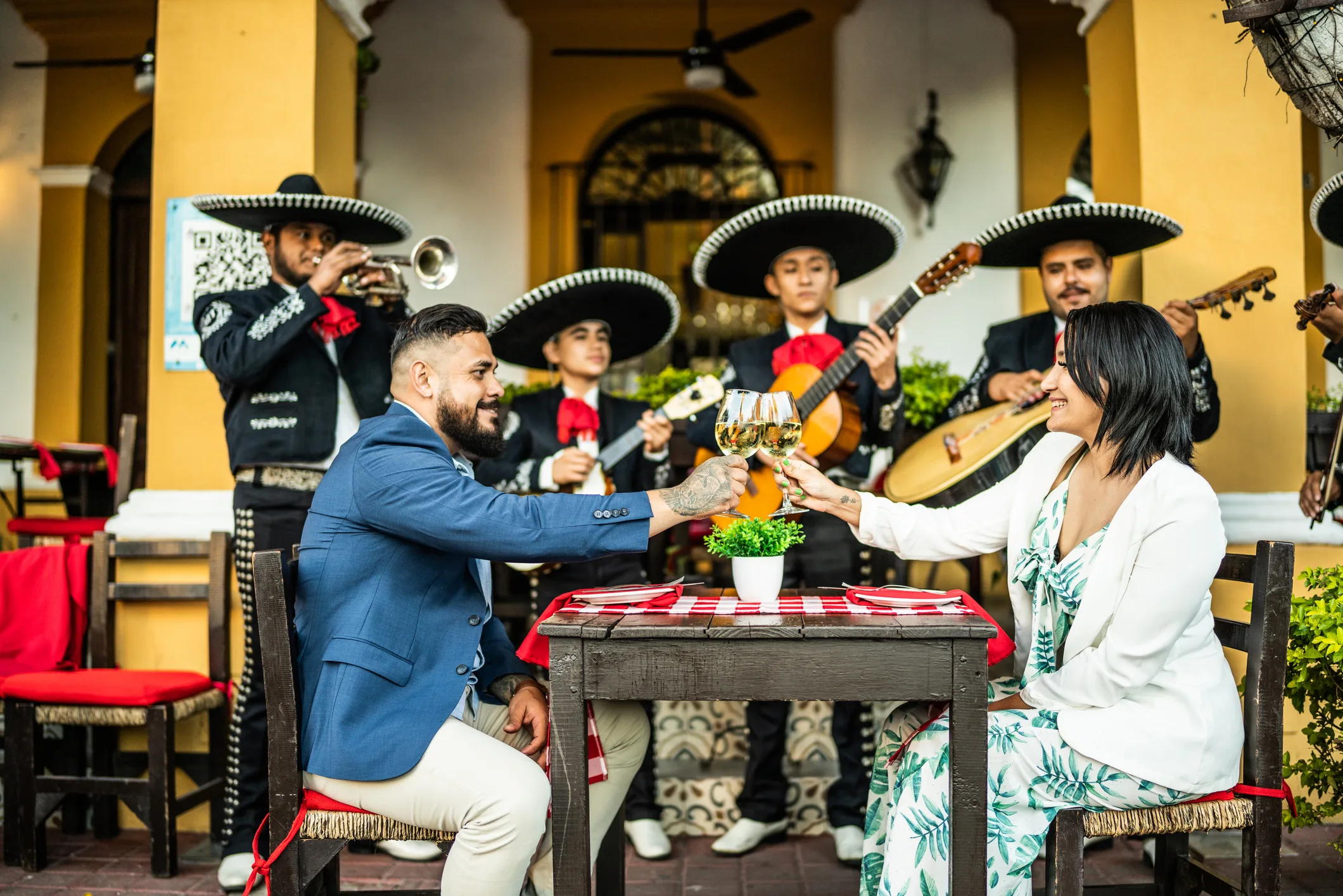 Ein Paar stößt mit Gläsern an, während Mariachi-Musiker im Hintergrund spielen.