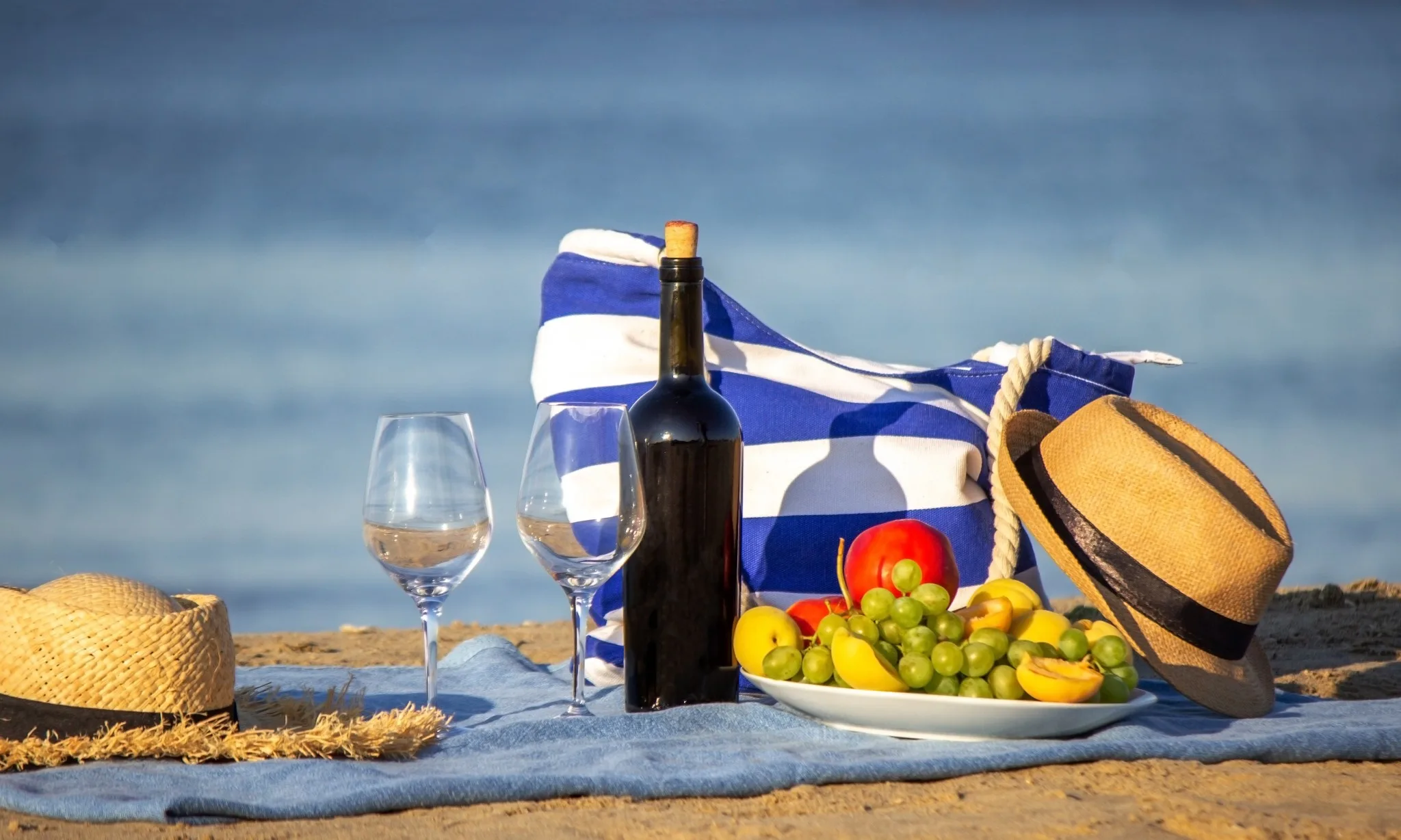 Ein Picknick am Strand mit Wein, Obst und Hüten auf einer Decke.