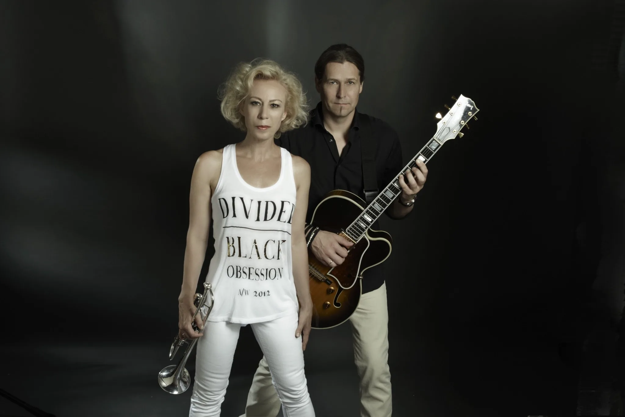 A musician duo poses against a dark background. The woman holds a trumpet, and the man holds a guitar.