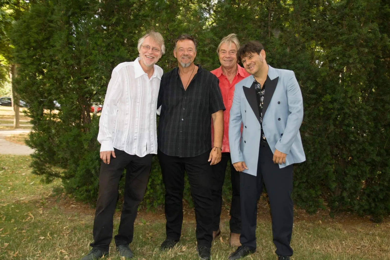 Four men smiling outdoors in front of green trees.